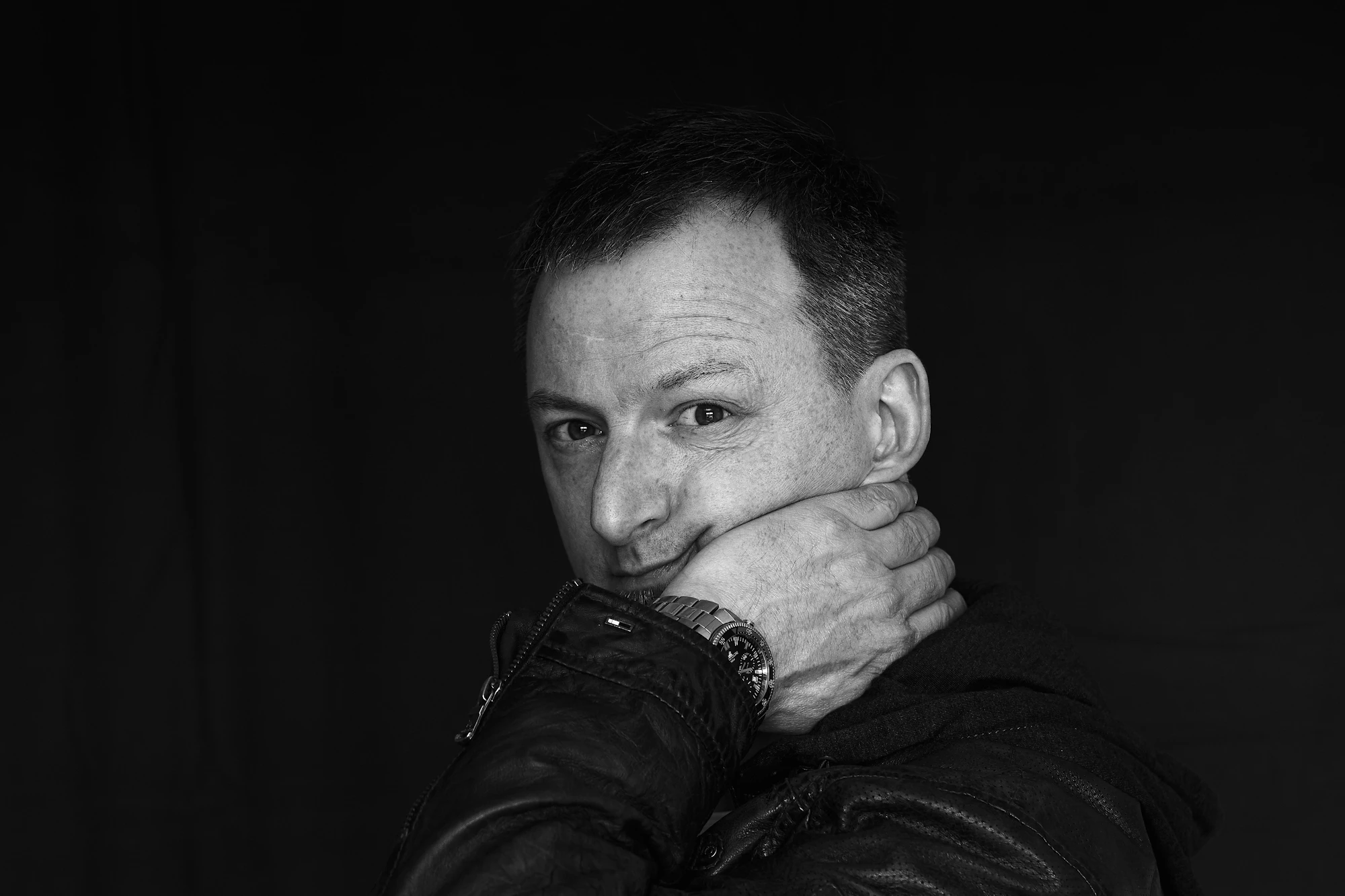 Ein Mann in einer schwarzen Lederjacke vor einem schwarzen Hintergrund. Der Mann schaut zum Betrachter und hat seine linke Hand unterm Kinn.