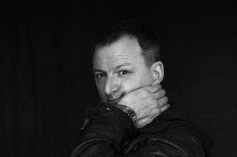 Ein Mann in einer schwarzen Lederjacke vor einem schwarzen Hintergrund. Der Mann schaut zum Betrachter und hat seine linke Hand unterm Kinn.