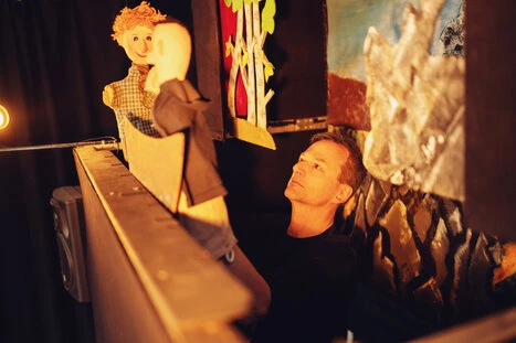 Ein Puppenspieler hinter seiner Puppenbühne.er hat zwei Figuren auf den Händen. Die Szene ist von oben links aufgenommen.