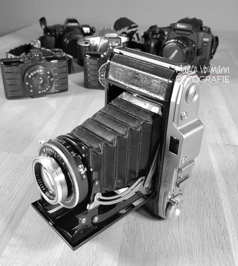 Eine aufgeklappte alte analoge Faltbalg-Kamera steht vor einer Anzahl anderer Kameras